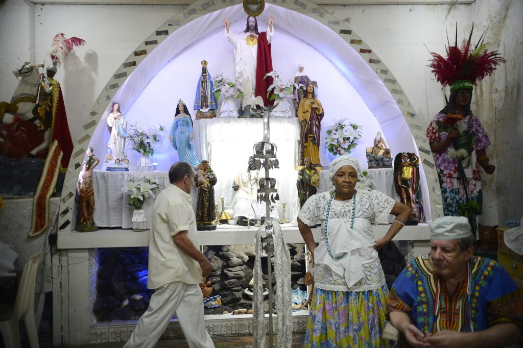 A photograph inside a traditional Umbanda terreiro, showing an altar with Catholic and Afro-Brazilian spiritual figures. An older woman dressed in white and colorful skirts stands near the altar, a man in ritual clothing sits to the side, and another person walks across the room. The space reflects the syncretic nature of Umbanda, with statues, flowers, candles, and ritual elements.